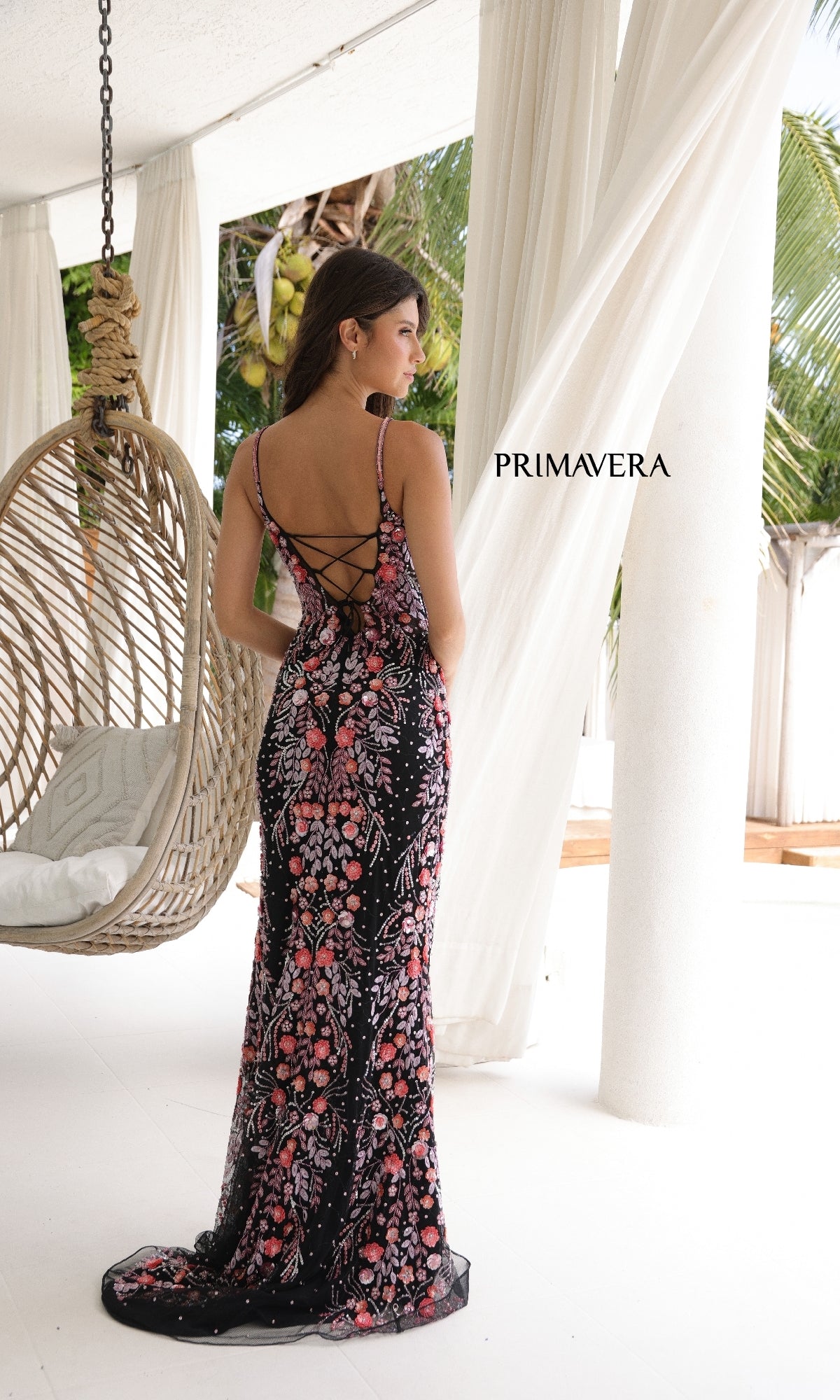 A woman in a black, floor-length, floral Primavera 4313 Lace-Up Long Beaded Prom Dress stands indoors near a hanging chair and white curtains.
